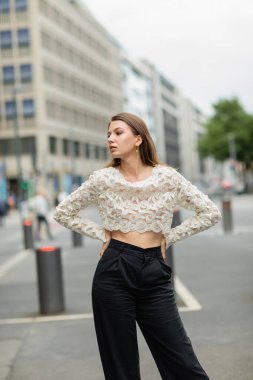 Almanya, Berlin 'de sokakta dantelli bluz ve yüksek bel pantolon giymiş modaya uyan genç bir kadın.