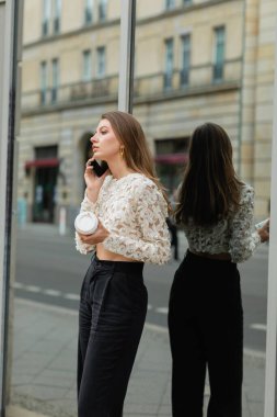 Modaya uygun genç bir kadın elinde kahve tutarak Berlin, Almanya 'da akıllı telefonla konuşuyor. 