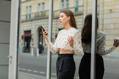 Berlin, Almanya 'daki bir binanın yakınına gitmek için akıllı telefon kullanan ve elinde kahve tutan trend bir genç kadın. 