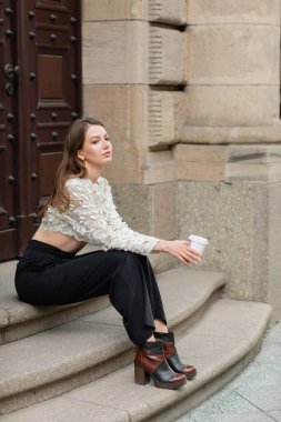 pretty young woman holding coffee to go while sitting on stairs near door in Berlin, Germany  clipart