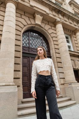 elegant young woman in lace top and high waist pants looking away on urban street in Berlin clipart