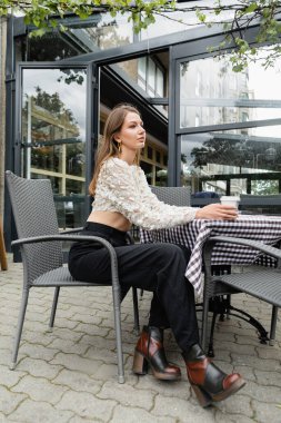 Berlin 'deki kafe yakınlarındaki terasta otururken elinde kahve tutan modaya uygun genç bir kadın.