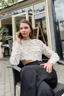 Stylish woman talking on smartphone while relaxing on terrace of cafe on street in Berlin clipart