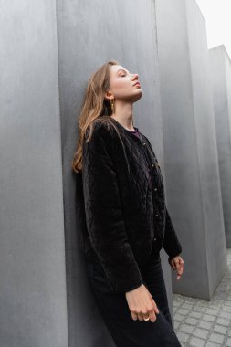 Relaxed young woman in jacket standing near Memorial to Murdered Jews of Europe at daytime in Berlin clipart