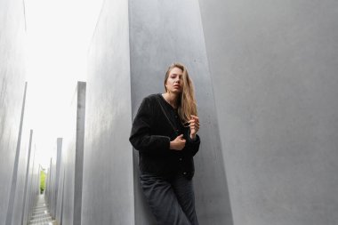 trendy young woman in jacket standing between Memorial to Murdered Jews of Europe in Berlin clipart