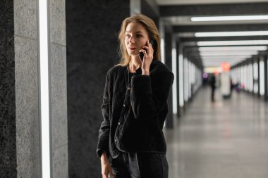 Modern binanın içinde dikilirken akıllı telefondan konuşan siyah ceketli şık bir kadın.