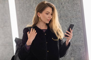 Siyah ceketli genç bir kadın sırt çantası tutuyor ve floresan lambaların yanında akıllı telefon kullanıyor.