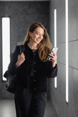 Sırt çantalı mutlu kadın floresan lambaların yanında akıllı telefon kullanıyor. 