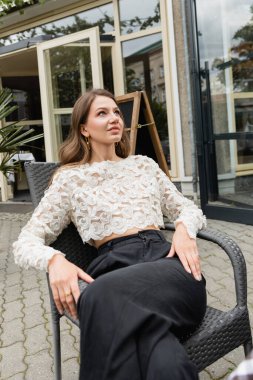 Gülümseyen ve rahat, dantelli, yüksek bel pantolonlu genç bir kadın sandalyede otururken gözlerini kaçırıyor.