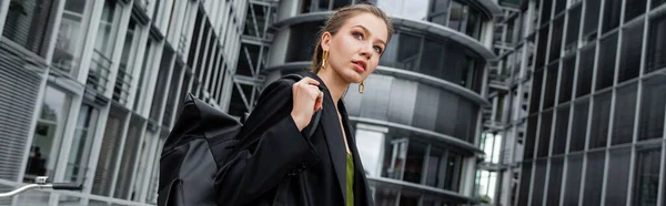 stock image young and fair haired woman in black jacket holding backpack and looking away, banner  