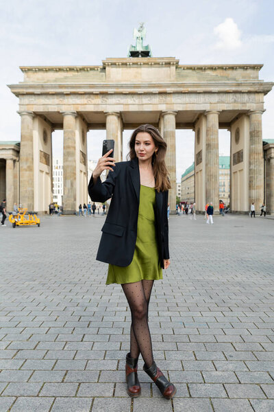 BERLIN, GERMANY - MAY 13, 2022: woman in blazer and silk dress taking selfie near Brandenburg Gate 