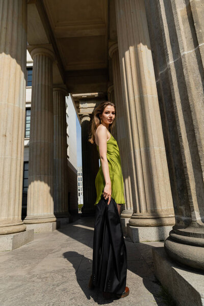 Elegant young woman in silk dress holding black jacket and looking at camera in Berlin, Germany 