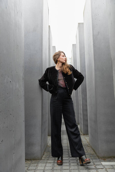 Full length of stylish young woman in black jacket and pants holding hands on hips in Berlin