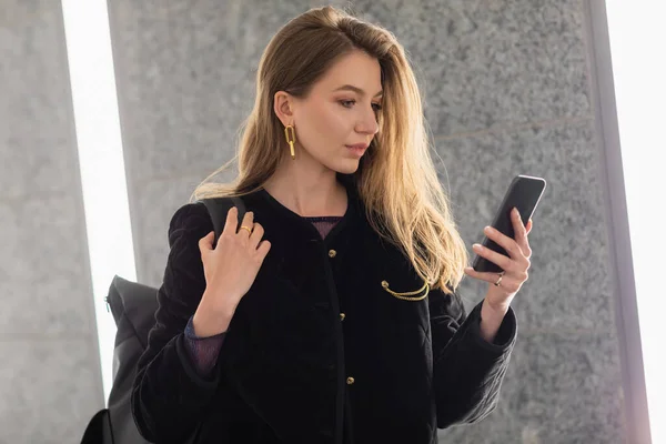 stock image young woman in black jacket holding backpack and using smartphone near lighting of fluorescent lamps