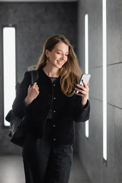 happy woman in jacket holding backpack and using smartphone near lighting of fluorescent lamps