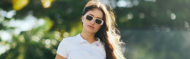 charming and young woman with brunette long hair standing in white polo shirt and trendy sunglasses near blurred and green palm trees in Miami, sunny day, travel, vacation, banner, sun-kissed  clipart