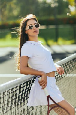 tennis court in Miami, sporty young woman with brunette long hair standing in white outfit and sunglasses while holding racket near tennis net, blurred background, iconic city, looking at camera  clipart