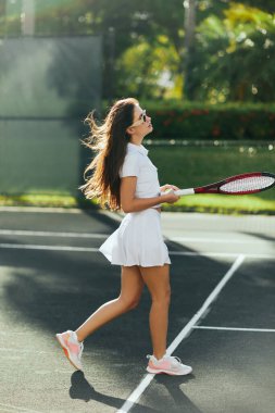 Uzun esmer saçlı atletik bir genç kadın spor beyaz kıyafetiyle yürüyor ve elinde raketle Miami, Florida 'da tenis kortunda, güneşli bir günde arka planda bulanık palmiye ağaçları tutuyor. 