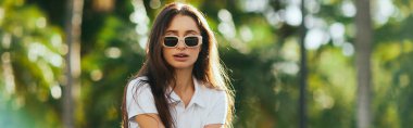 captivating young woman with long hair standing in white polo shirt and trendy sunglasses near blurred and green palm trees in Miami, sunny day, travel, vacation, banner, iconic city  clipart