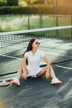 Miami 'de tenis kortu, esmer, uzun saçlı, beyaz elbiseli, güneş gözlüklü, raket ve topun yanında dinlenen, arka planda bulanıklık olan, yaz mevsimi,