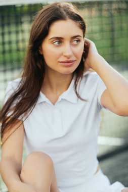 portrait of attractive young woman wearing white polo shirt and looking away while adjusting long brunette hair after training on tennis court, tennis net on blurred background, Miami, Florida  clipart