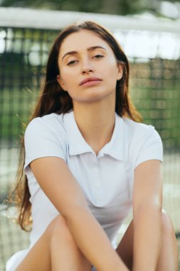 Esmer, uzun saçlı, beyaz polo tişörtlü ve tenis kortunda antrenmandan sonra kameraya bakan kadın tenisçinin portresi bulanık arka planda tenis ağı, Miami, Florida