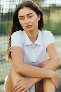 Miami 'de tenis kortu, esmer, uzun saçlı, beyaz polo tişörtlü ve antrenmandan sonra kameraya bakan kadın tenisçi portresi, bulanık arka planda tenis ağı, Florida