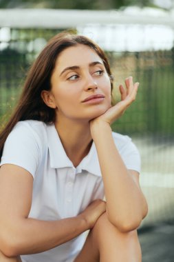 tennis court in Miami, portrait of dreamy female tennis player with brunette hair wearing white polo shirt and looking away after training, tennis net on blurred background, Florida clipart