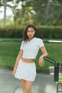 summer lifestyle, sporty young woman with brunette hair standing in white outfit with skirt and polo shirt near cart with balls, blurred background, sun-kissed, tennis court in Miami  clipart