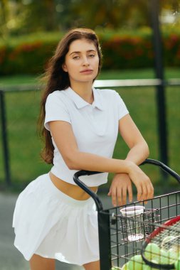 healthy habits, sporty young woman with brunette hair standing in white outfit with skirt and polo shirt and leaning on cart with balls and racket, blurred background, tennis court in Miami  clipart