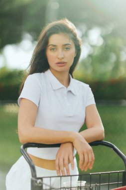 tennis court in Miami, sporty young woman with brunette hair standing in white polo shirt near tennis cart, blurred green background, looking at camera, pretty tennis player, soft filter clipart