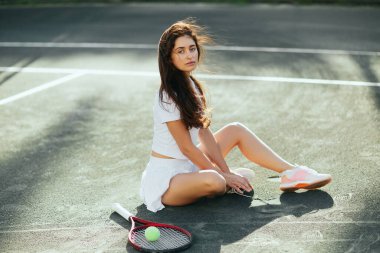 Kadın maç üstüne dinleniyor, uzun saçlı bayan tenisçi beyaz kıyafetli, raketin yanında oturuyor, top asfaltta, arka plan bulanık, Miami, tenis kortu, dinlenme zamanı, gölgeler, güneşli bir gün.