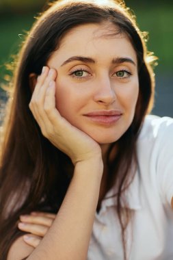portrait of delightful young woman with brunette long hair posing in white polo shirt and looking at camera, blurred background, Miami, Florida, iconic city, natural makeup, laid-back  clipart