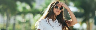 summer vibes, pretty woman adjusting long brunette hair and standing in white polo shirt and sunglasses on urban street in Miami, blurred background, palm trees, vacation in Florida clipart