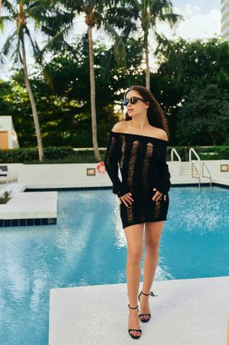 Siyahi örgü elbiseli, güneş gözlüklü çekici kadın Miami 'de palmiye ağaçlarına karşı parıldayan suyla birlikte açık havada yüzme havuzunun yanında poz veriyor. 