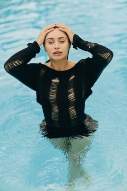 no makeup look, attractive and sexy brunette woman in black knitted outfit posing inside of outdoor swimming pool during vacation in Miami, alluring, luxury resort, Florida  clipart