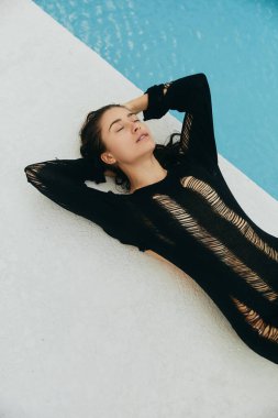 Lüks otel, siyah örgü elbiseli seksi bir kadın Miami 'de parlayan suyla yüzme havuzunun yanında yatıyor, yaz tatili, gençlik, havuz kenarında rahatlama, rahat duruş, üst manzara.
