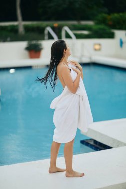 young brunette woman with wet hair wrapped in white towel standing next to outdoor swimming pool with shimmering water in Miami, summer getaway, youth, poolside relaxation, vacation mode  clipart