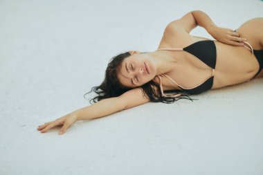Siyah bikinili çekici bir kadın, ıslak saçlı, lüks otellerde poz veren, Miami, Florida, ABD, bulanık arka plan, beyaz yüzeye uzanmış, havuz kenarında dinlenme 