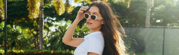 очаровательная и молодая брюнетка с длинными волосами, стоящая в белой футболке и модных солнцезащитных очках рядом с размытыми и зелеными пальмами в Майами, солнечный день, путешествия, отдых, баннер 