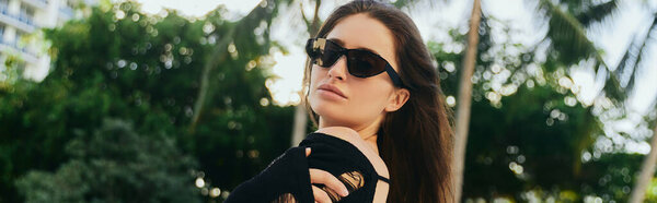 summer getaway, sexy brunette woman with tanned skin, tourist in black knitted dress and sunglasses looking at camera in luxury resort during vacation in Miami, green palm trees, banner 