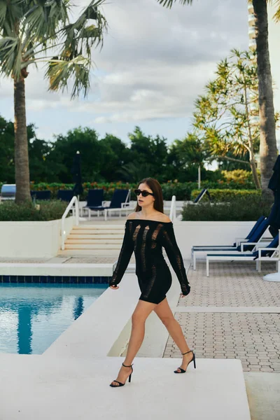 在迈阿密 穿着黑色针织服装 戴着太阳镜 穿着高跟鞋 头戴一双迷人的女人 站在户外游泳池旁边 在棕榈树下闪烁着光芒的水 在豪华度假胜地 在日光浴床上度假 — 图库照片
