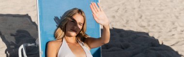 Genç, güzel sarışın bir kadın Miami sahilinde sörf tahtasının yanında duruyor. Tuzlu esintiyi hissediyor..