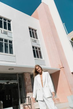 Beyaz takım elbiseli, çarpıcı sarışın bir kadın Miami 'de modern bir binanın önünde duruyor..