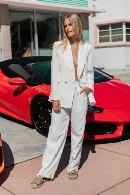 Şık, genç, sarışın bir kadın hareketli Miami şehrinde canlı kırmızı bir spor arabanın yanında güvenle duruyor..