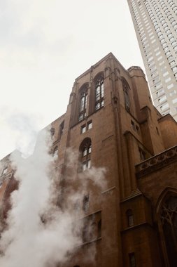 New York 'ta buhar ve gökdelenin yanındaki kırmızı tuğlalı katolik kilisesinin alçak açılı görüntüsü
