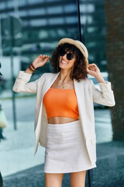 A stylish woman in a white blazer poses in front of a glass building in Europe. clipart