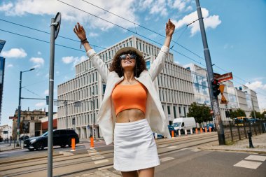 A woman in stylish attire enjoys a sunny day in Europe, arms raised in delight. clipart