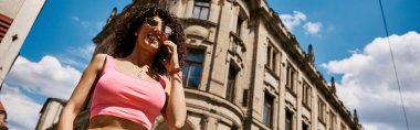 A woman in stylish attire smiles brightly as she walks through a European city on a sunny day. clipart