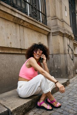 A woman in pink and white outfit sits on a step in a European city, talking on her phone. clipart